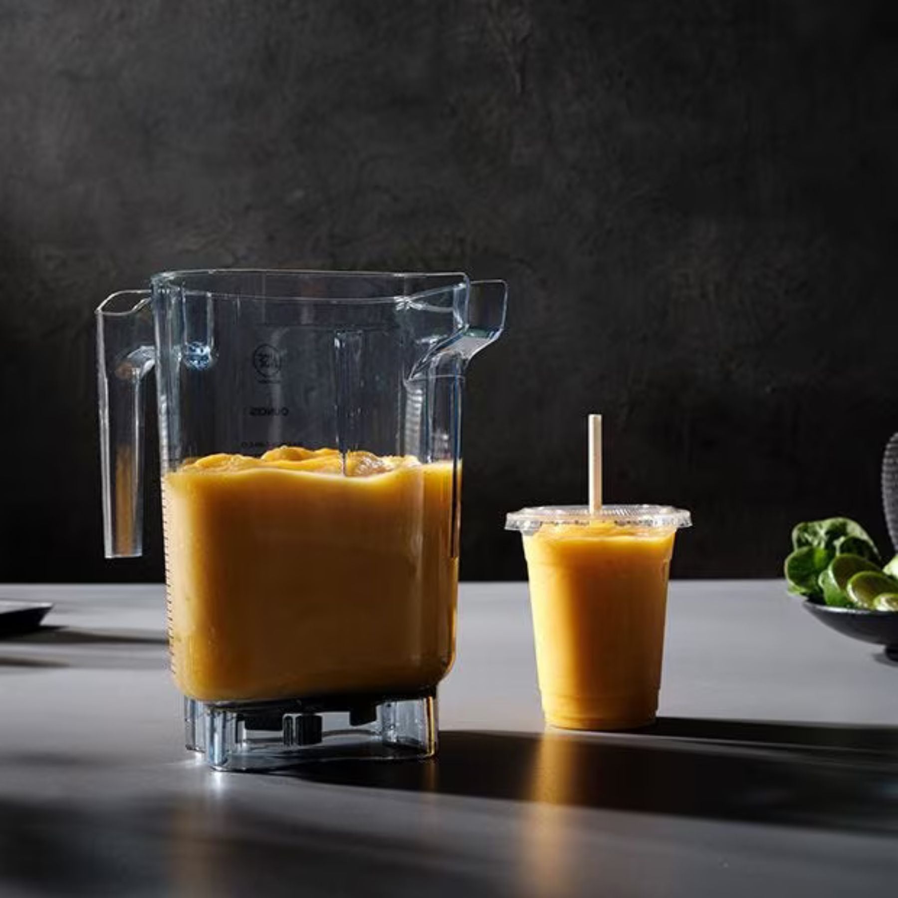 Blender with a yellow smoothie and a cup of smoothie on a dark surface.