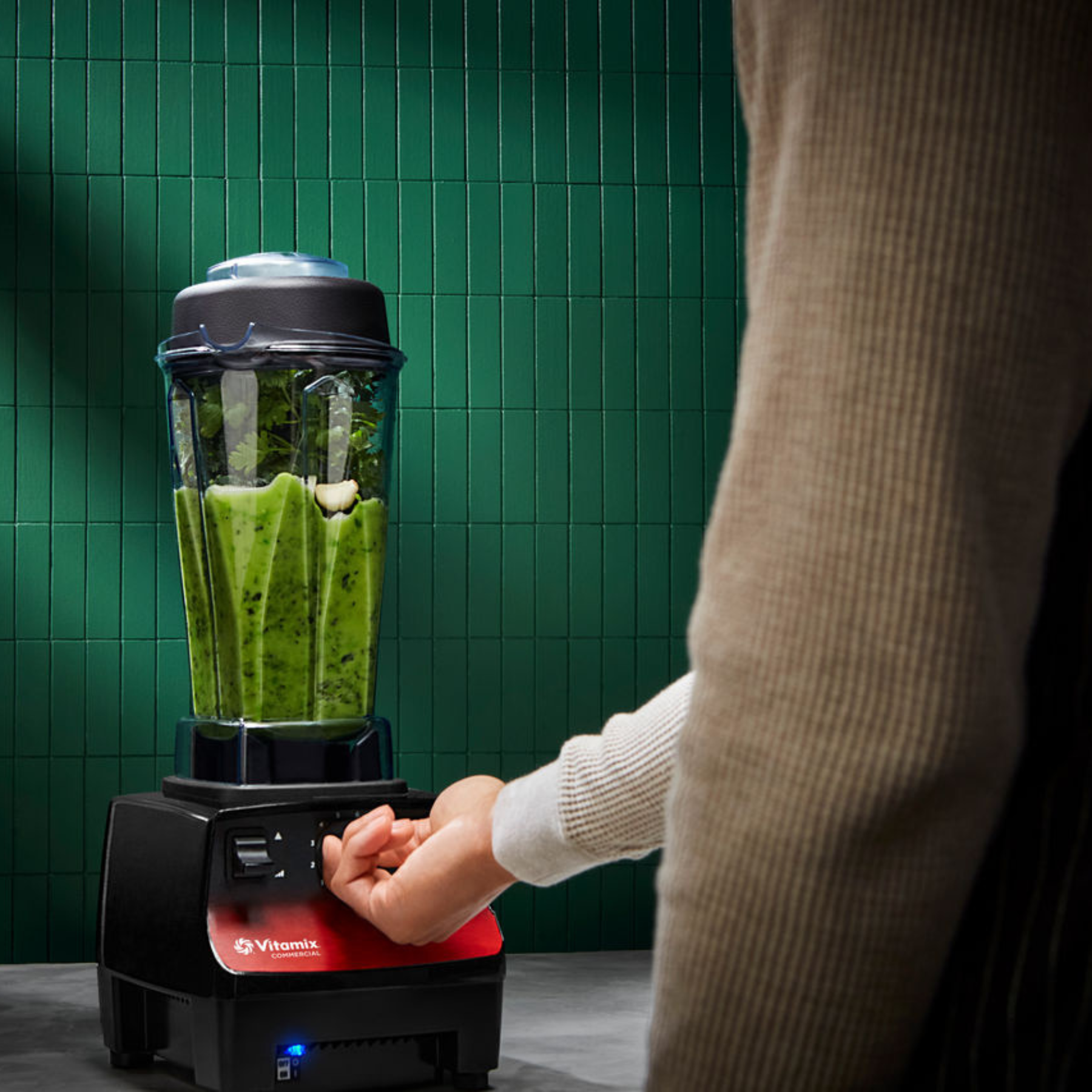 Person operating a Vitamix Vita-Prep 3 | Blenders Direct with a green smoothie inside against a green tiled wall.