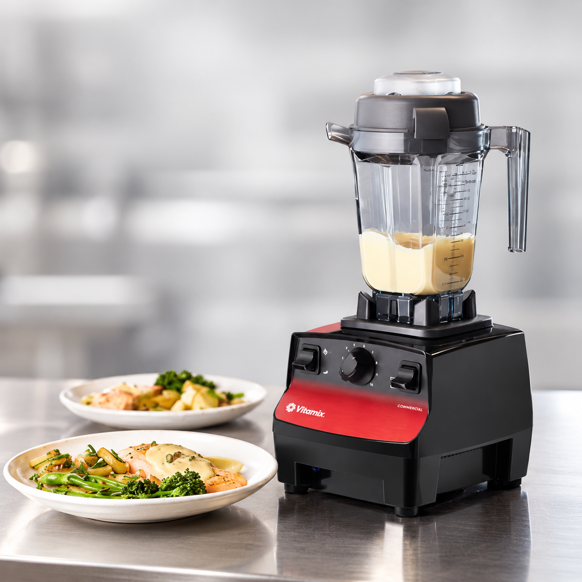 A Vitamix professional blender with a clear jar containing a pale yellow liquid, set on a stainless steel countertop next to two white plates of food in a blurred kitchen environment.